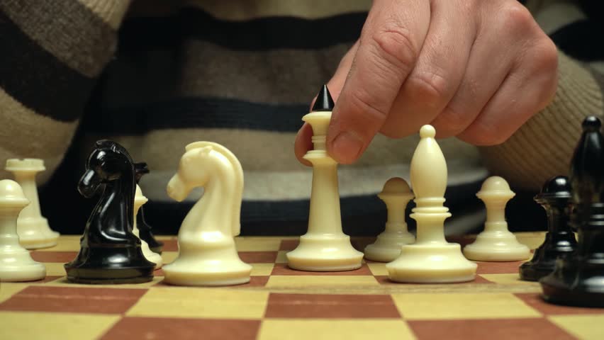Hand of a player positions a white queen on a chessboard, surrounded by black and white chess pieces, highlighting the strategic gameplay in a warm, inviting indoor environment