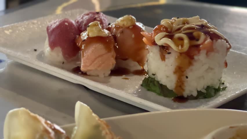 Handheld clip of a sushi platter with tuna and salmon nigiri and a drizzled roll on a rectangular plate, with a water glass and gyoza on the table.