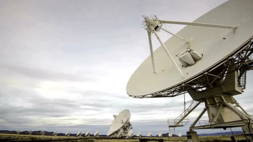 Radio telescope array dishes angled toward sky in high desert landscape tracking signals and communications while clouds gather overhead on quiet afternoon