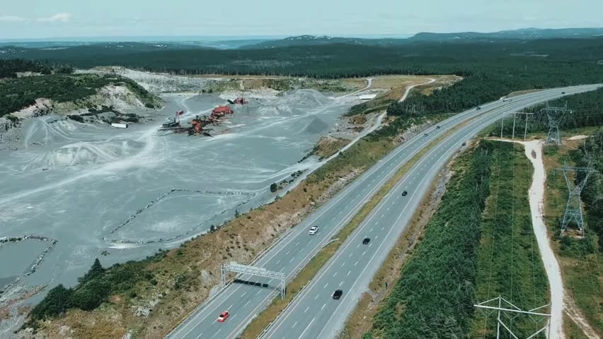 Cars drive on highway next to large quarry in Newfoundland, Canada. View includes surrounding landscape and power lines.