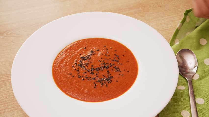 Hand placing fresh green basil leaf as garnish on top of hot red tomato and pepper cream soup in white ceramic bowl. Food styling for cooking vlog.
