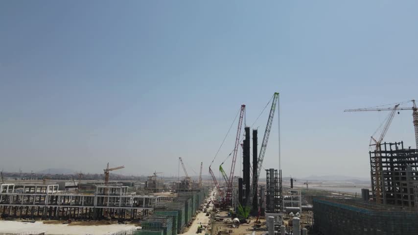 Aerial view reveals large industrial complex under construction. Cranes erect steel framework. Buildings and other equipment spread across the vast site on a clear day in China.