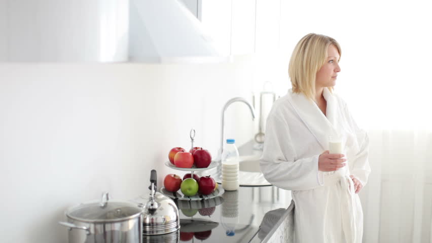 Woman in bathrobe standing in kitchen and drinking milk
