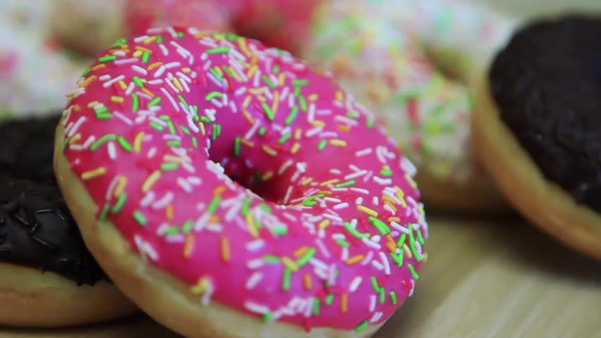 Close up view displays pink frosted donut with colorful sprinkles. Delicious looking baked pastry on table with chocolate frosted donuts in background.