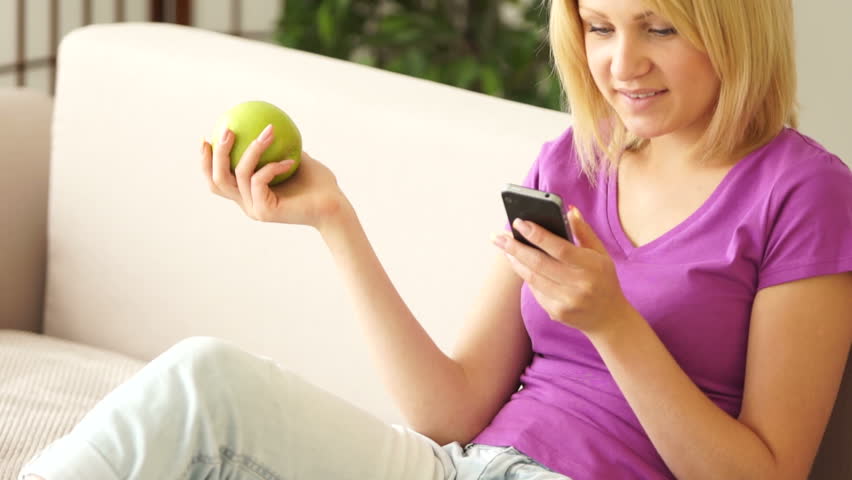Woman sitting on sofa with phone and eating an apple
