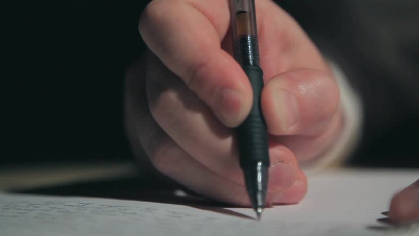 Close-up of a hand gripping a pen writing on paper in low light. The focus on the pen movement and paper captures an authentic moment of writing and creativity.