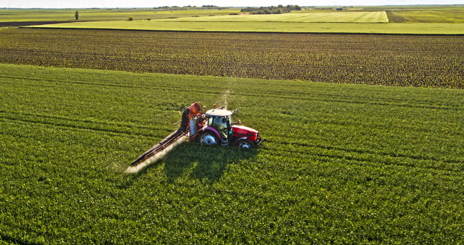 Farming tractor spraying a vast green field, performing pest control on a farm at sunset