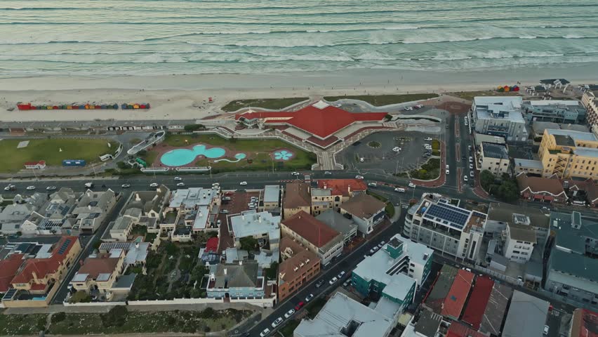 Aerial view looks down on coastal complex with red roof and swimming pool beside beach, while ocean waves and golden hour light shape sunset atmosphere in Muizenberg, Cape Town, South Africa.