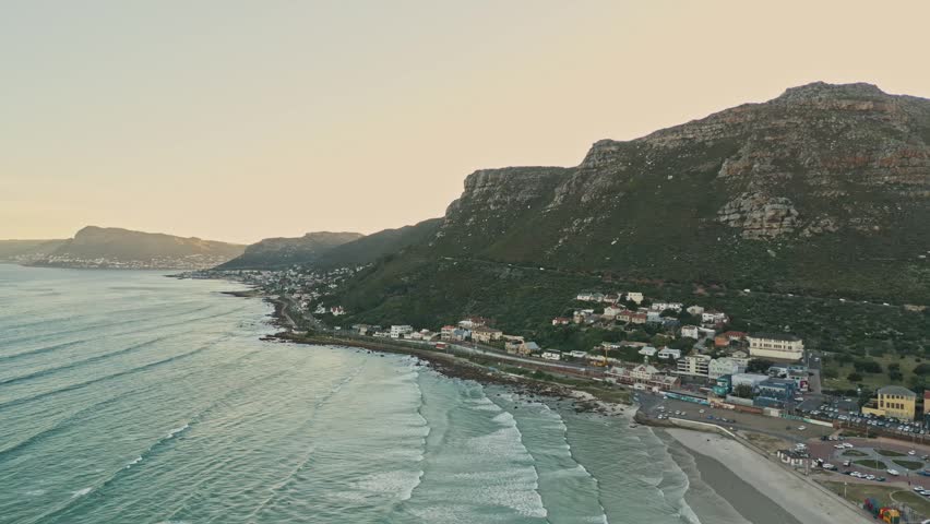 Aerial drone view follows long shoreline beside low-rise coastal town and mountain slope, while golden hour sunlight warms ocean, rooftops, and beach at sunset in Cape Town, South Africa.