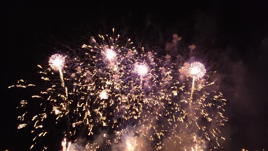 A stunning cinematic shot featuring massive, sequential explosions of professional fireworks in the night sky. Radiant purple circular patterns and cascading golden sparks illuminate the horizon with a magical glow, creating a festive and celebratory atmosphere. Perfect for projects related to celebrations, New Year