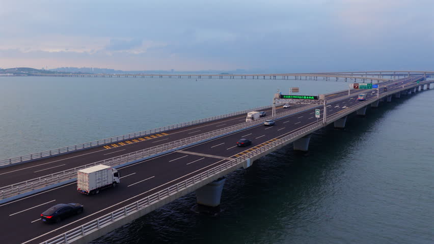 Blue hour aerial scenery of Jiaozhou Bay Bridge Qingdao, multi-level sea interchange highway, freight lorries with shipping containers traveling over sea, modern coastal transport engineering in China