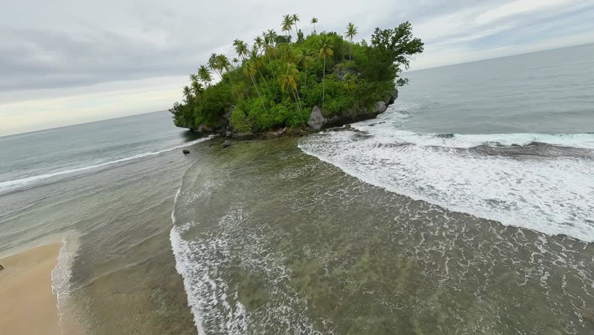 FPV drone flying over a small tropical islet surrounded by ocean waves, rising above palm trees to reveal a breathtaking coastal coastline view