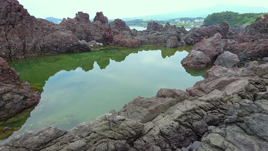 Camera pans across jagged volcanic rock formations to reveal calm green water pool at Socheonji Reservoir on Jeju Island Korea