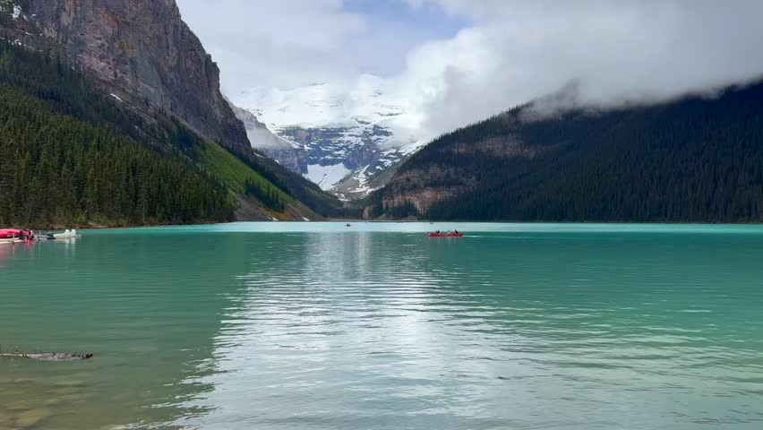 Beautiful view on the Lake Louise in National Park of Banff, Canada. Two kayakers navigate a serene lake, flanked by towering mountains and lush greenery, with snow-capped peaks in the distance and cloudy skies above