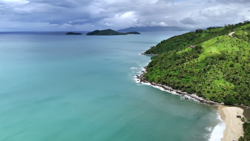 Aerial drone view of a turquoise coastline with a dense jungle headland and grassy hillside, two forested offshore islands under a dramatic stormy sky with surf on rocks, Taytay, Palawan, Philippines.