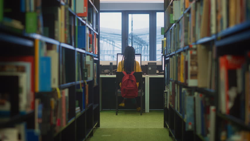 University Library Study: Black Female Student Sits at Desk, Uses Desktop Computer for Online Research and Class Assignment. Back View Camera Moves Between Rows of Bookshelves, Classmates Walk Past