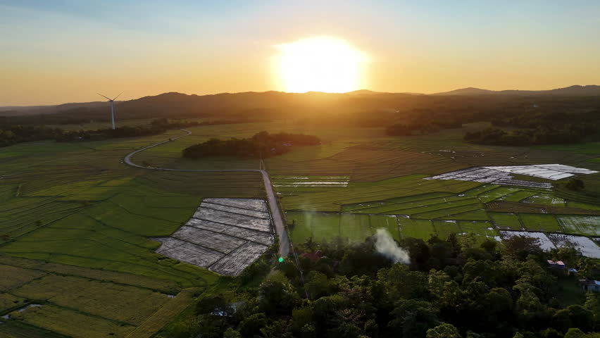 Aerial drone view of a wind turbine beside a rural road cutting through flat patchwork green and fallow rice fields at golden sunset, San Lorenzo Wind Farm, Guimaras, Philippines.
