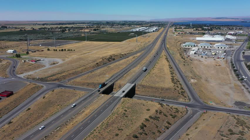 4K aerial drone footage of Interstate I-84 freeway near Boardman, Oregon, USA on a hot midsummer day during the dry season. Static aerial view showing light traffic, highway overpass, intersections, entrance and exit ramps, surrounding farmland and dry landscape near the industrial area of Boardman. Transportation infrastructure connecting eastern Oregon with the Columbia River region, featuring passenger vehicles and commercial trucks traveling through the rural high desert landscape