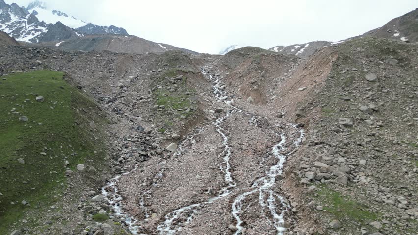 Aerial drone footage of a crystal-clear glacier-fed mountain stream flowing through the alpine landscape on the hiking route to Titov Lake and the Peak of Memory in Kazakhstan. Rocky terrain, snow-covered peaks and untouched wilderness highlight the beauty of the Tien Shan mountains.