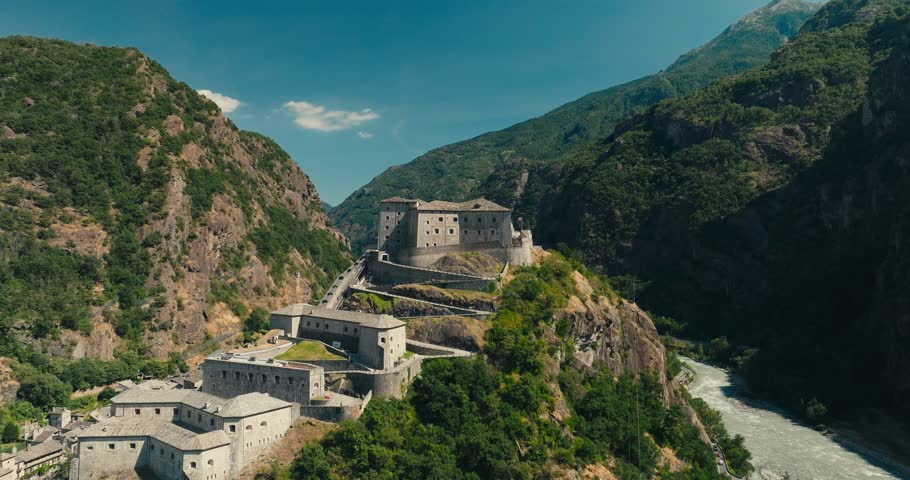 Drone rises above historic Fort Bard, revealing the winding river and deep Aosta Valley framed by lush green mountains beneath a clear blue summer sky in northern Italy.