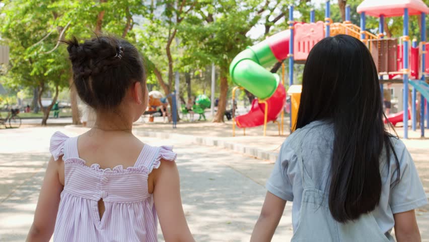 Two young girls hold hands while running toward a colorful playground in Lumpini Park, Thailand.