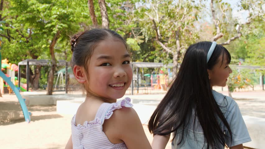 Two happy young girls hold hands and run through a sunny playground in Lumpini Park.