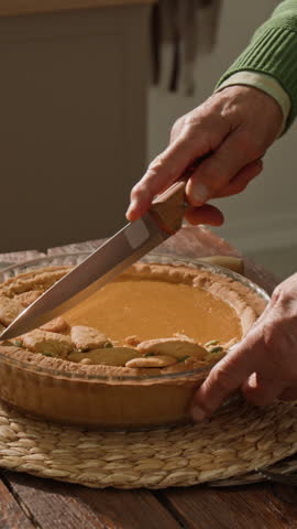 Vertical shot of unrecognizable senior man cutting homemade pumpkin pie with knife. Footage is ideal for Thanksgiving marketing campaigns and autumn holiday food promotions