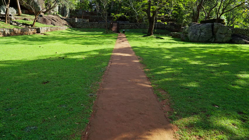 A red earth path crosses bright green lawns between rocks and shady trees in the boulder gardens of the ancient Sigiriya fortress in Sri Lanka.