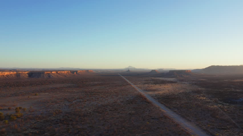 Slow panoramic drone pan of Ugab Valley, Namibia at sunset. Straight road crosses vast plain, geological formations visible. Clear cloudless sky