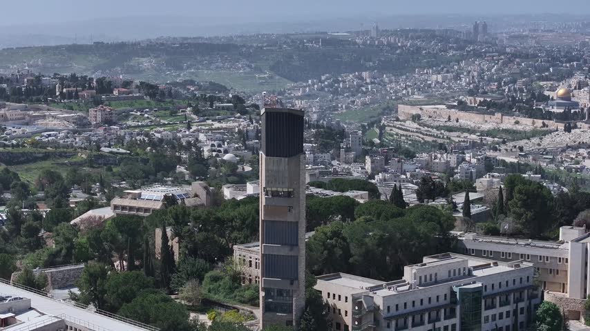 Drone shot moving up near Hebrew University campus tower revealing Mount of Olives and surrounding hills of Jerusalem on a bright sunny day.