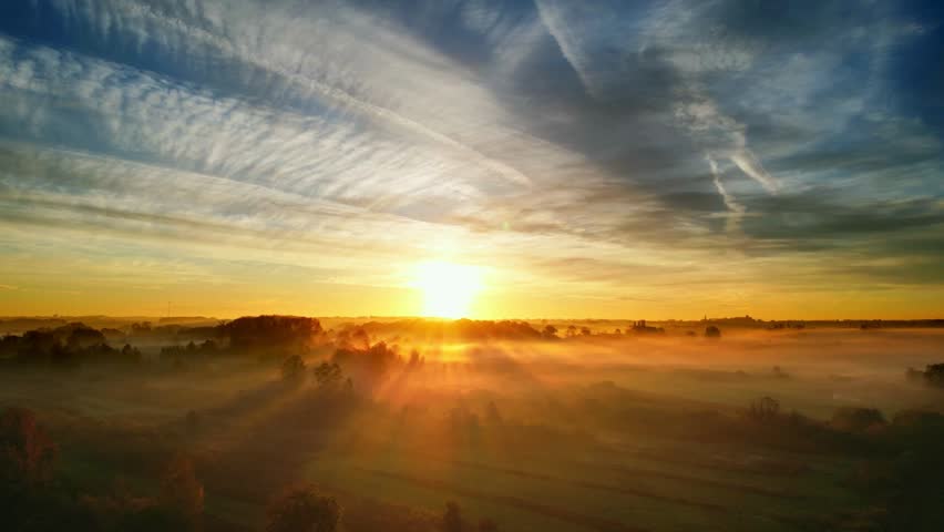 A breathtaking sunrise paints the sky with golden hues, casting light over a misty, rural landscape with unique cloud formations.