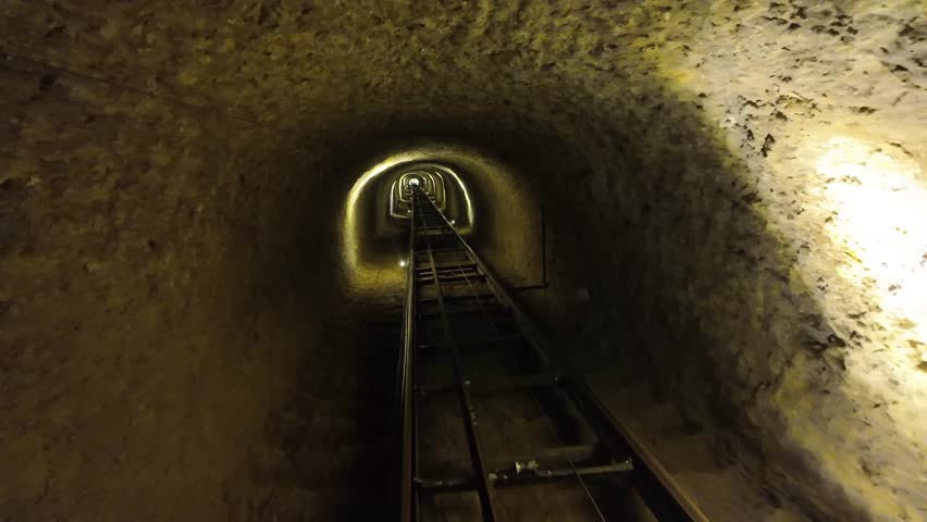 A long, illuminated underground tunnel with metal tracks stretches into the dark distance, creating a mysterious perspective.

