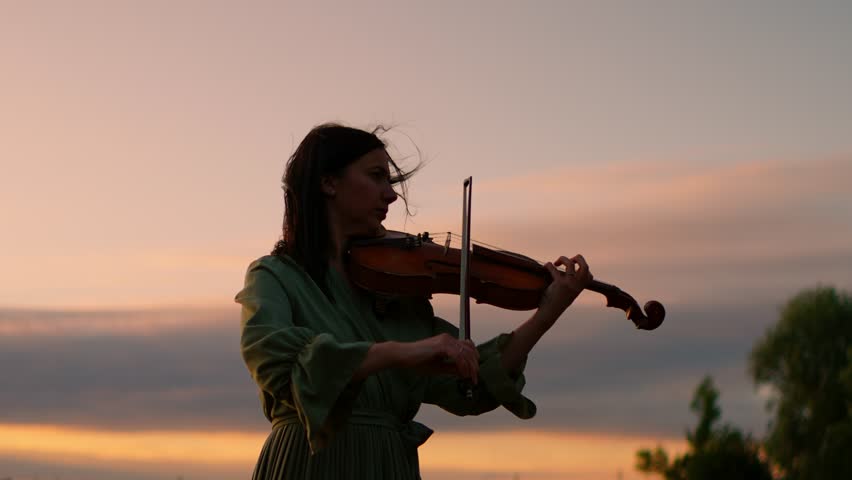 Silhouette of a woman playing violin outdoors at tranquil sunset, graceful musician in flowing dress performing emotional melody against colorful evening sky
