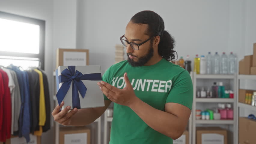 Man wearing volunteer tshirt holding a wrapped gift box in a donation center room with shelves and clothing racks; community support gratitude.