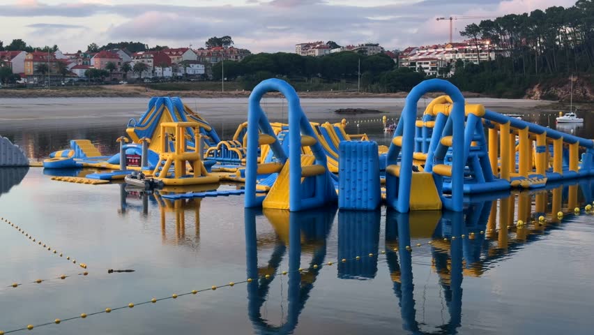 Blue and yellow inflatable floating water park on calm water beside a coastal town at dusk