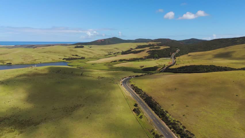 Car drives along curving highway through green rolling hills in Northland, New Zealand, small freshwater lake and distant blue coastline spread across grazing farmland. Aerial drone wide shot