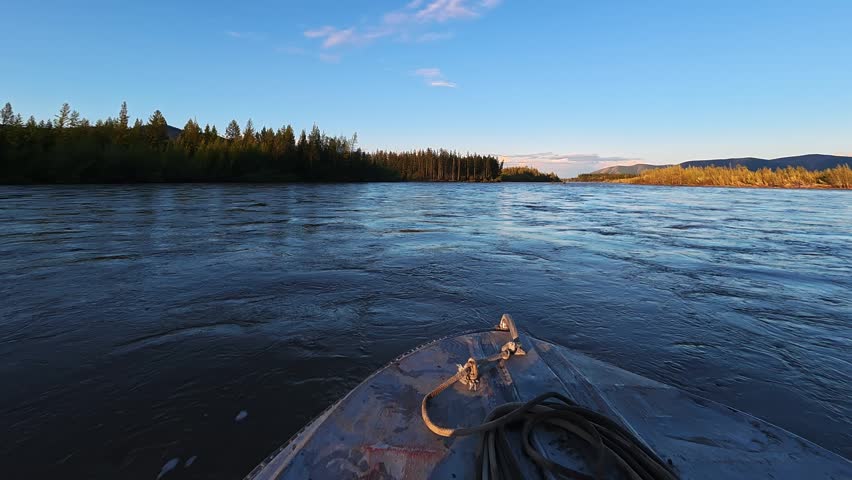 Front view from a small boat navigating calm river waters, surrounded by dense forests and rolling hills under a clear blue sky during golden hour.