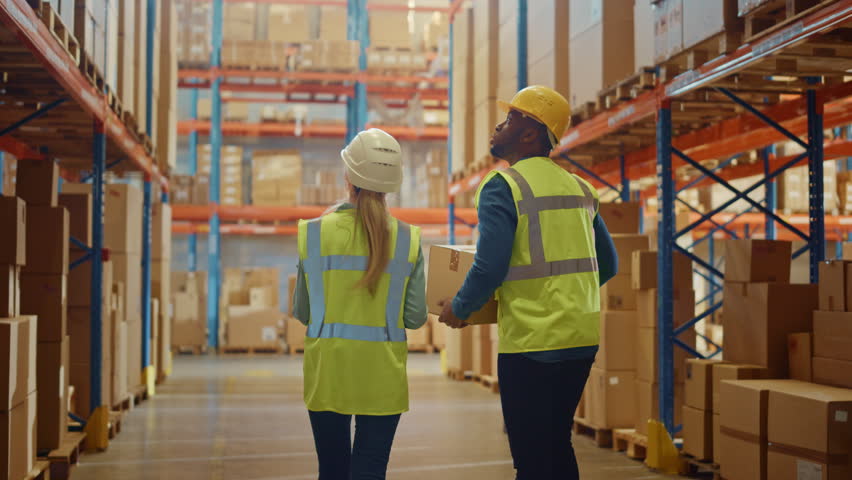 Online Retail Fulfillment Warehouse: Black Male Employee Holds Cardboard Parcel and Chats With Blonde Caucasian Female Coworker While Walking Between Storage Racks. Back View, Slow Motion