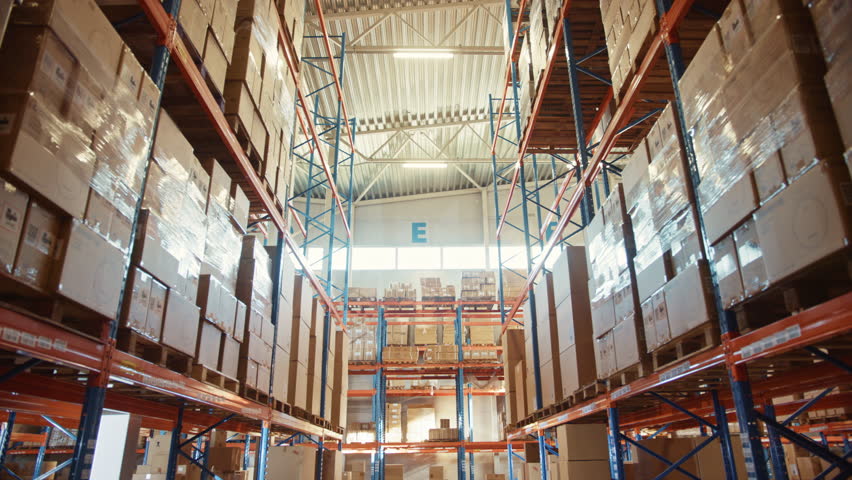 E-commerce Online Retailer Distribution Center Warehouse With Tall Rows of Shelves Holding Cardboard Boxes, Packages and Consumer Products Prepared for Customer Orders. Descending Camera View in Aisle