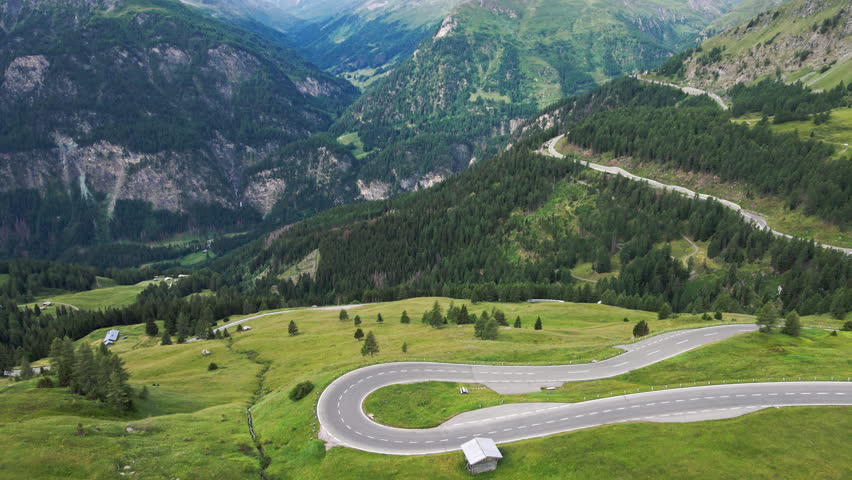 Grossglockner High Alpine Road winds across green mountain slopes in Austria on a summer day. Aerial view of sharp hairpin turns above a forested valley in the Alps.