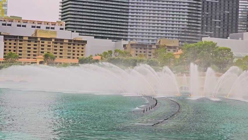 Bellagio Fountains daytime performance with tall sprays, mist, rippled turquoise water, resort buildings, stone arches, and Las Vegas Strip skyline in a scenic travel shot.