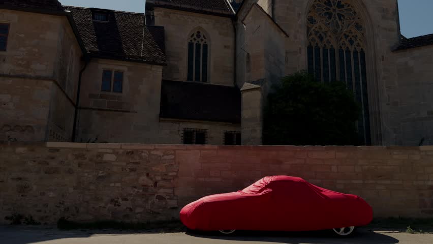 Contemporary lifestyle scene with a covered sports car parked behind a historic church in Germany, blending modern living, personal belongings and traditional European architecture.