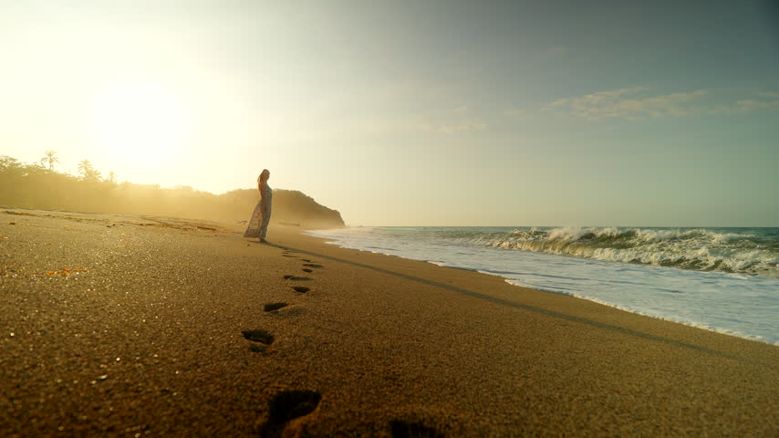 Footprints on remote beach with female traveller near waves crashing at sunrise