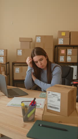 Woman counting cash with hands beside cardboard boxes and laptop in a packing area inside a building; small business focus.