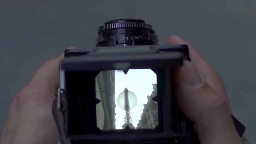 Cinematic close-up of a photographer using a vintage film camera to capture the Eiffel Tower in Paris, France. The iconic landmark appears framed through the camera viewfinder between historic Parisian buildings, creating a nostalgic travel photography concept. Atmospheric urban scene representing tourism, European culture, travel memories, photography, Paris architecture, and visual storytelling, ideal for editorial, documentary, travel, and creative media projects.