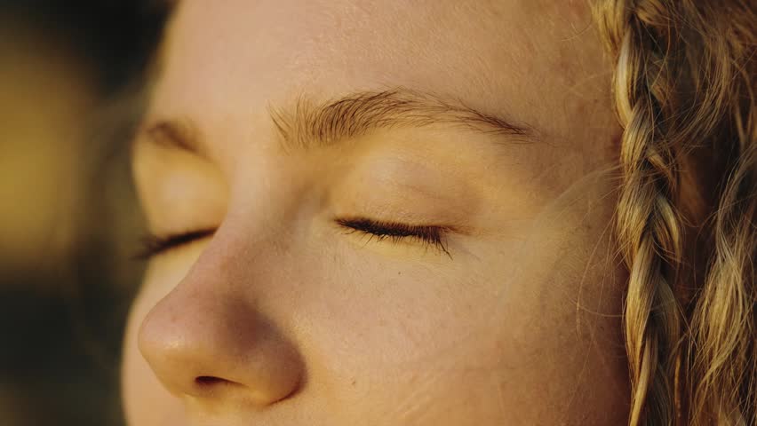 Stunning close-up shot of a young woman with gorgeous green eyes opening and closing them in golden sunset light. Braided blonde hair, tranquil mood, Ibiza coastline background, natural beauty.