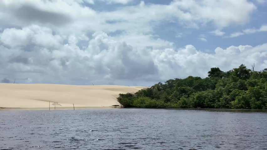 Low-angle shot from a boat cruising the Preguiça River, featuring large coastal sand dunes and wind turbines under a cloudy sky in Maranhão, Brazil.
