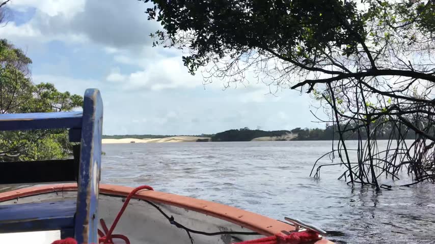 POV shot from the bow of a wooden boat cruising along the Preguiça River with lush green tree-lined shores under a cloudy sky in Maranhão, Brazil.