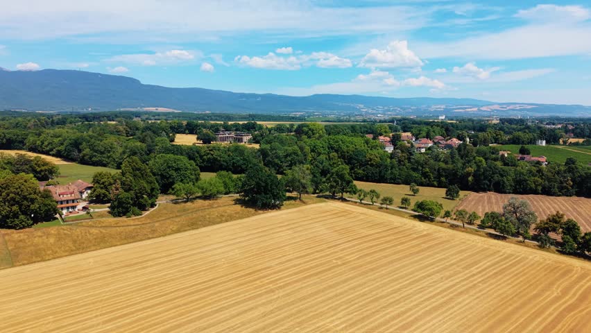 Aerial view of the historic Domaine de Belle-Ferme surrounded by golden wheat fields and lush green trees under a bright blue sky in Bogis-Bossey, Vaud, Switzerland.