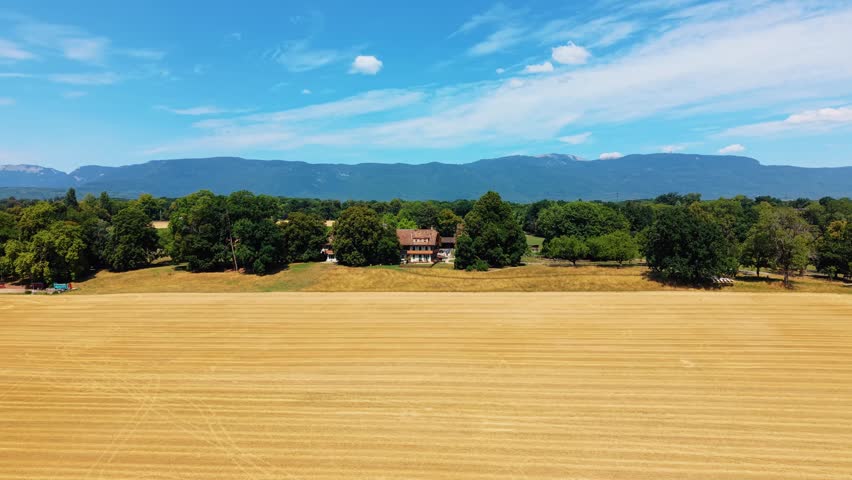 Aerial view of Domaine de Belle-Ferme surrounded by golden wheat fields and lush trees with mountains in the distance in Bogis-Bossey, Vaud, Switzerland.